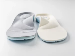 Aangepaste podiatrische inlegzolen naast standaard steunzolen op wit klinisch oppervlak, met zichtbare gewelfondersteuning en ergonomische vormgeving.