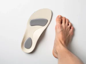 Orthopedische inlegzool naast een blote voet op wit oppervlak, bovenaanzicht met zichtbare gewelfde ondersteuning.
