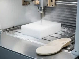 CNC-freesmachine snijdt op maat gemaakt orthopedisch inlegzool uit wit schuimblok, afgewerkte zool op stalen werkvlak.