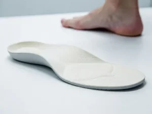 Orthopedische inlegzool op wit klinisch oppervlak met licht gebogen textuur, onscherpe blote voet op de achtergrond.