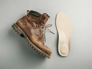 Versleten wandelschoen naast een orthopedische inlegzool op lichtgrijze ondergrond, bovenaanzicht met zacht natuurlijk licht.