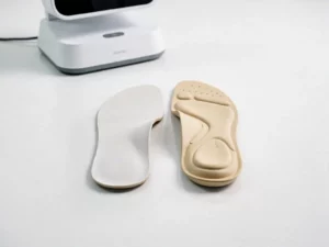 Twee op maat gemaakte orthopedische inlegzolen naast elkaar op wit klinisch oppervlak, met zichtbare gewelfondersteuning en 3D-scanner op achtergrond.