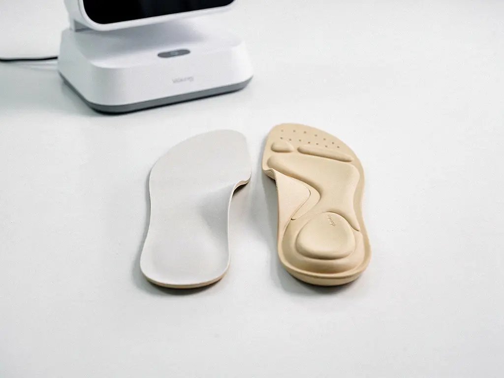 Twee op maat gemaakte orthopedische inlegzolen naast elkaar op wit klinisch oppervlak, met zichtbare gewelfondersteuning en 3D-scanner op achtergrond.