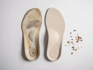 Versleten orthopedische inlegzool naast een nieuwe inlegzool op wit oppervlak, met zichtbaar verschil in schuimkwaliteit en kurrelkorrels.