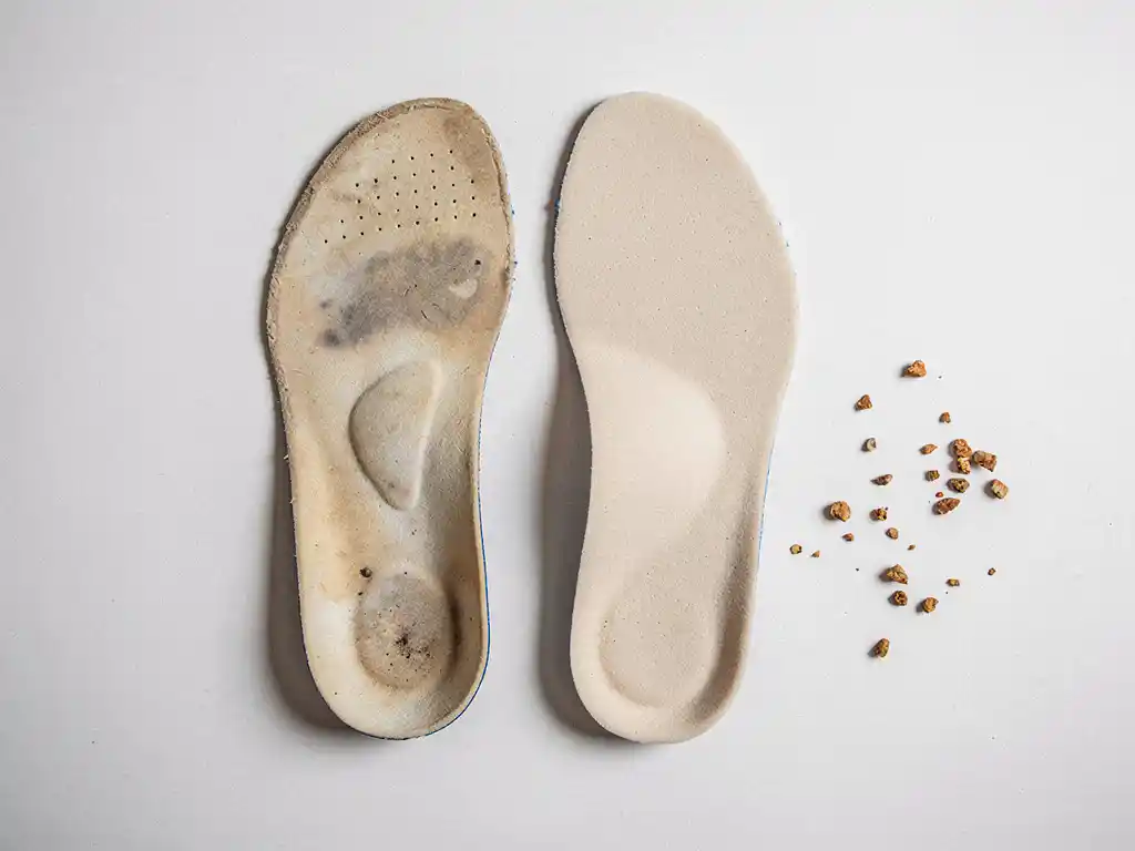 Versleten orthopedische inlegzool naast een nieuwe inlegzool op wit oppervlak, met zichtbaar verschil in schuimkwaliteit en kurrelkorrels.
